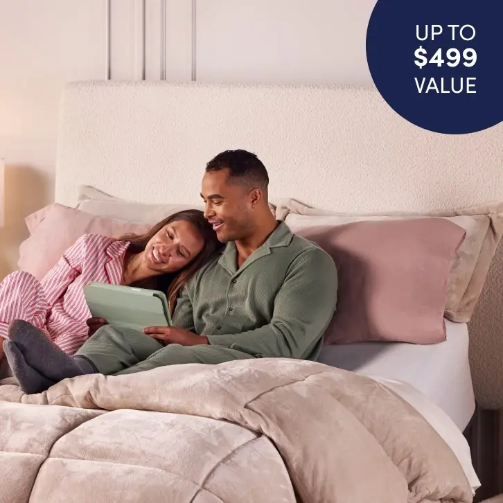 A smiling couple is sitting up in bed, wearing pajamas and enjoying content on a tablet together. The bed has soft beige and blush bedding with multiple pillows. A circular label in the top right corner states "UP TO $499 VALUE."