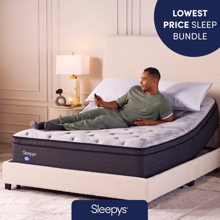 A man in green pajamas is relaxing on an adjustable bed while holding a remote control. The mattress is branded "Sleepy's" and features a gray quilted top. The head section of the bed is elevated, and there’s a modern nightstand with a lamp and decorative items beside the bed. A label in the top right corner reads "LOWEST PRICE SLEEP BUNDLE."